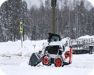 Compact white and black skid-steer loader with red wheels parked on snowy ground near a large snowbank and snow-covered trees.