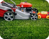 Close-up of a red and silver lawn mower on green grass next to a yellow and red toy lawn mower.