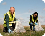 Two people wearing yellow safety vests and gloves picking up trash in a grassy field.
