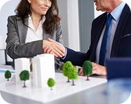 Two business professionals shaking hands over an architectural model of buildings and trees.