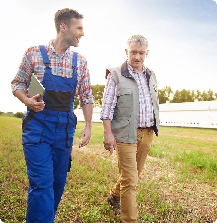 Two men walking and talking in a field, one in blue overalls holding a tablet, the other in a gray vest and brown pants.