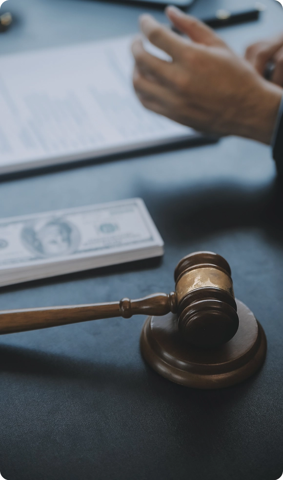 Wooden judge's gavel resting on a sound block with blurred hands, legal document, and stack of money in the background.