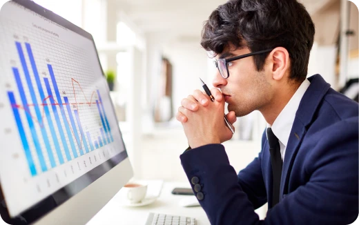 Man in a navy blazer and glasses intently analyzing bar and line charts on a desktop computer screen.