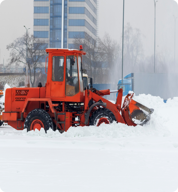 Orange snow plow clearing snow on a street with a blue truck and tall building in the background.