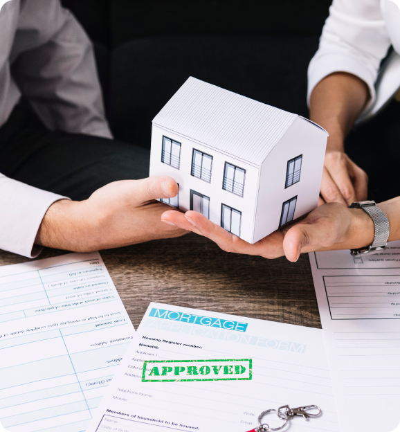 Two people holding a small white model house above mortgage approval documents on a table.