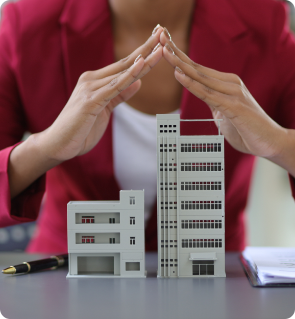 Person in a red jacket forming a protective gesture over two small architectural building models on a table.