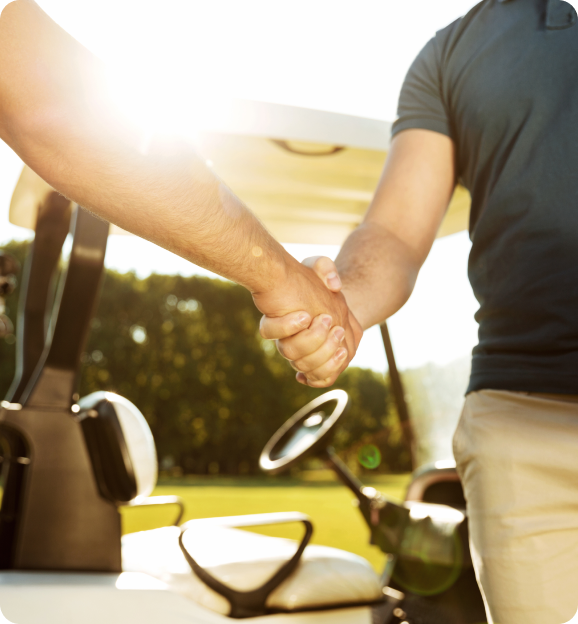 Two people shaking hands in front of a golf cart on a sunny day.