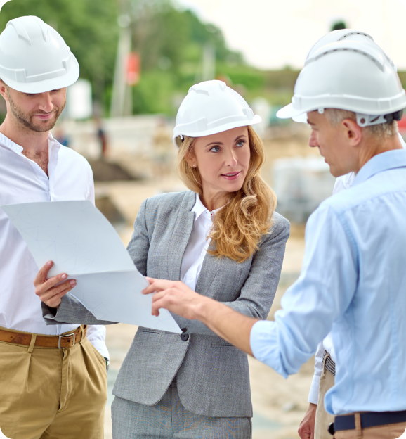 Three construction professionals wearing white hard hats reviewing documents at a construction site.