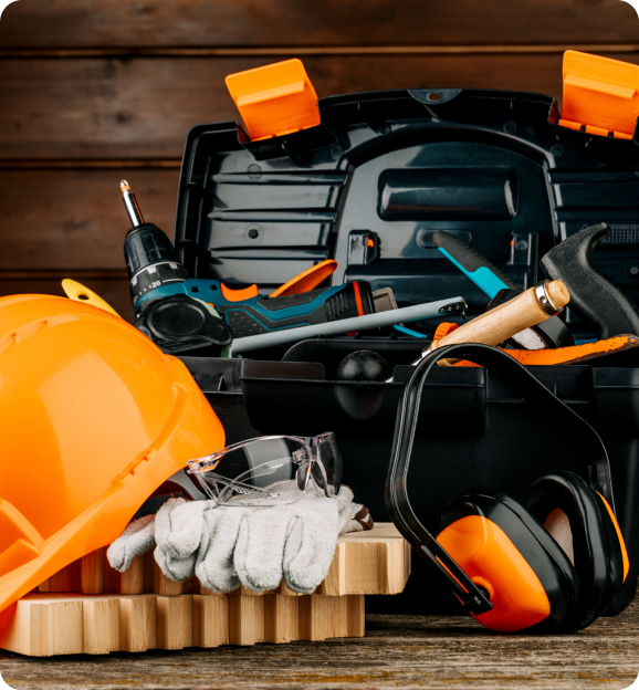 Open black toolbox with power drill, hammer, and other tools, next to a yellow hard hat, safety gloves, protective glasses, and orange earmuffs on a wooden surface.