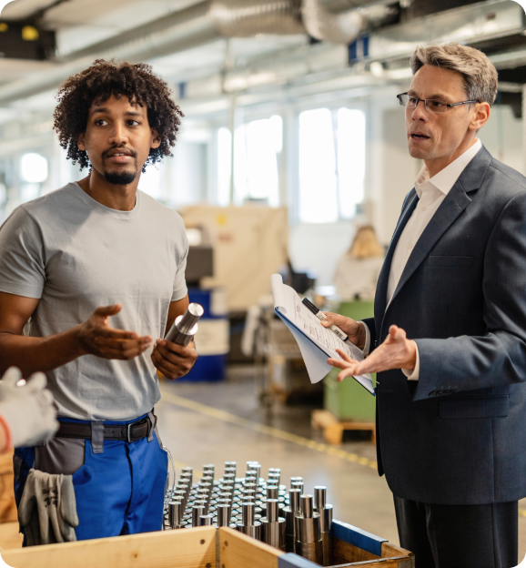 Two men in a factory discussing cylindrical metal parts with one holding a clipboard and the other holding a metal piece.
