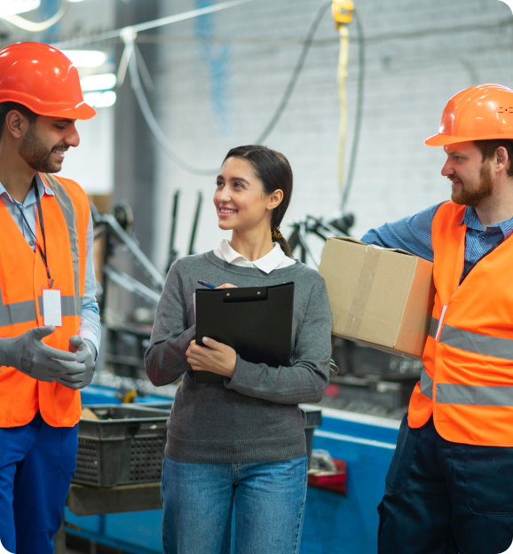 Two male workers in orange safety vests and helmets talking to a woman holding a clipboard in an industrial setting.