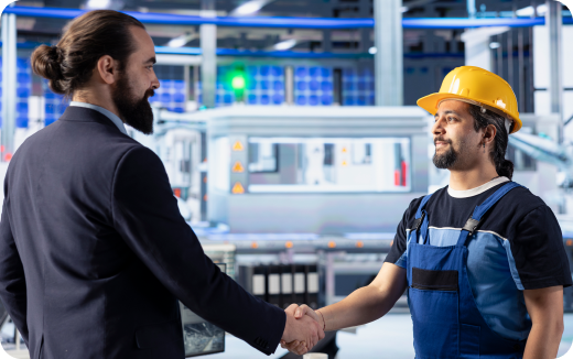 A businessman in a suit shaking hands with a factory worker wearing a yellow hard hat and blue overalls in an industrial setting.