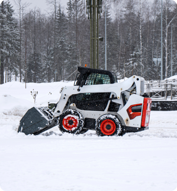 Compact white skid-steer loader with red wheels clearing snow in a snowy outdoor area with trees in the background.