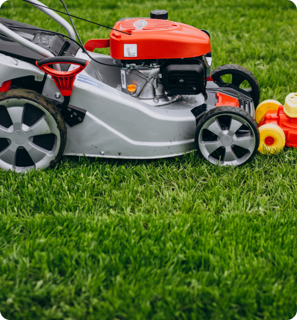 Close-up of a red and gray lawn mower on green grass with a toy lawn mower beside it.