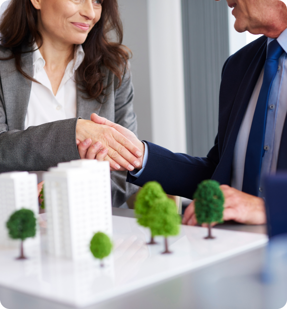 Two business professionals shaking hands over a table with a scale model of buildings and trees.