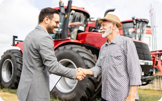 Man in a suit shaking hands with an older man wearing a straw hat and plaid shirt in front of red tractors.