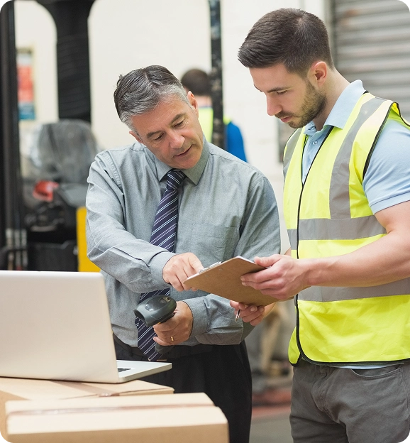 Warehouse supervisor instructing a worker in a safety vest while holding a barcode scanner and clipboard near boxes and a laptop.
