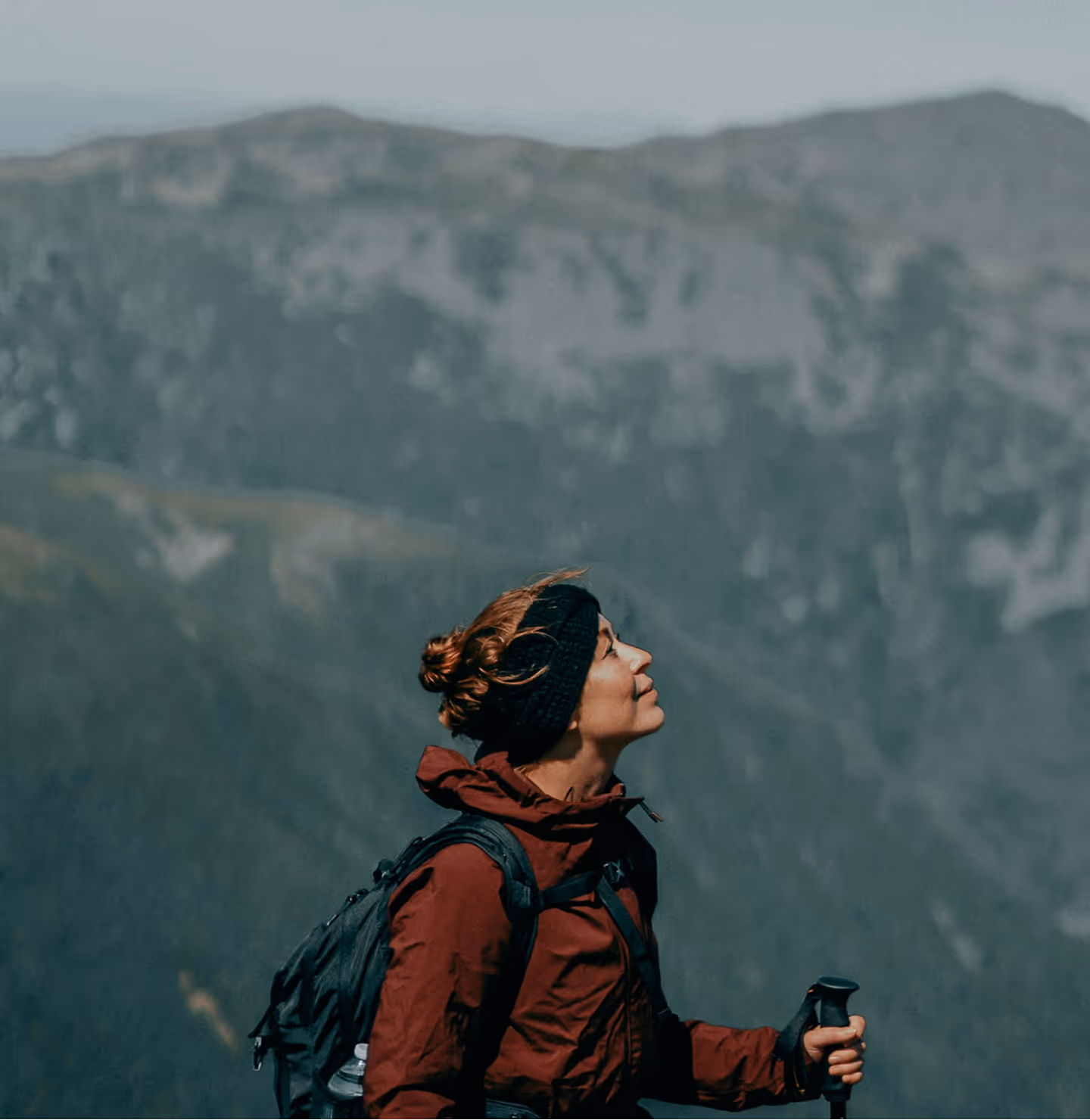 Woman in a red jacket and headband hiking with poles, mountains in the background.