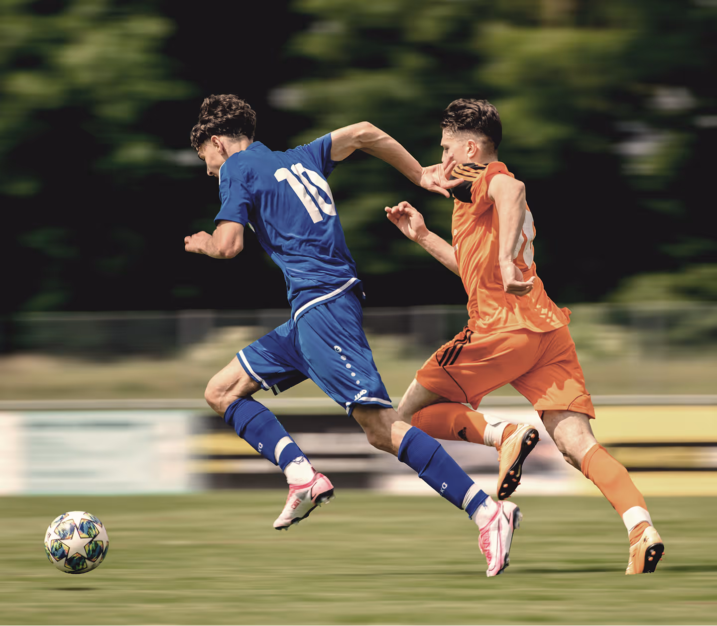 Two soccer players in blue and orange uniforms sprinting on a field chasing a ball.