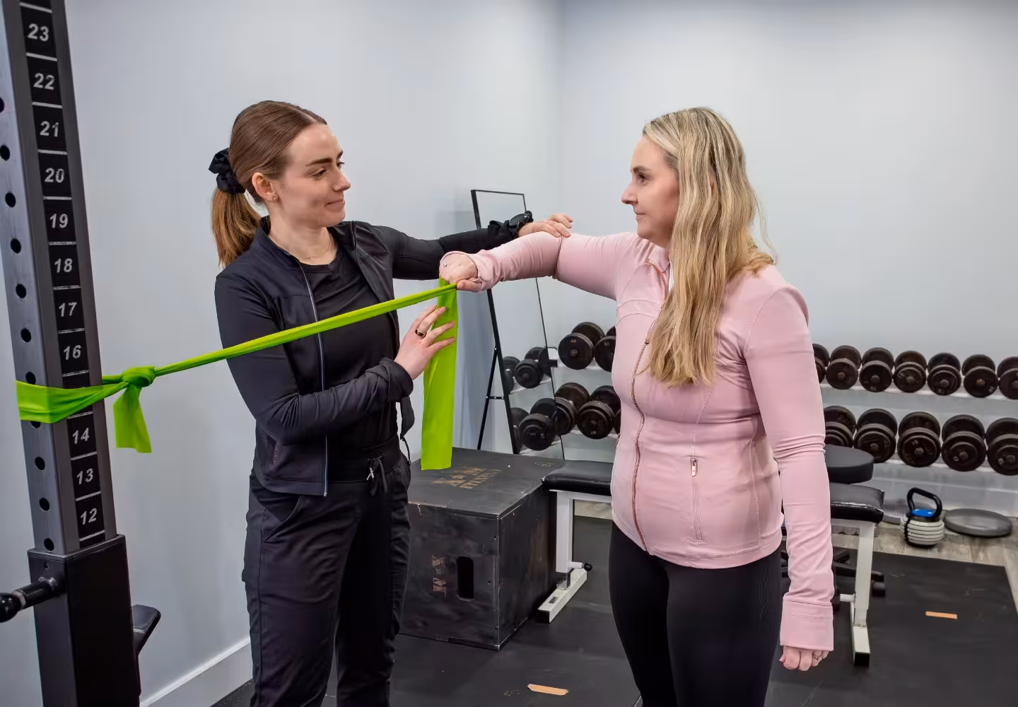 Physical therapist assists woman with arm exercise using green resistance band in gym.