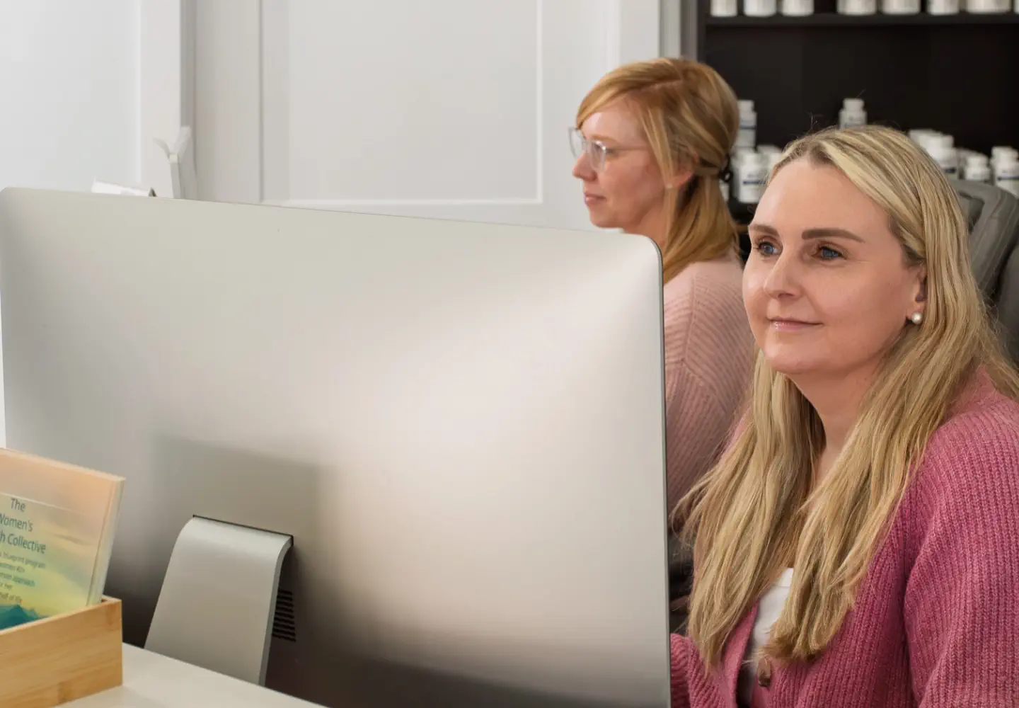Two women working at a desk with a large computer monitor and shelves of bottles behind them.