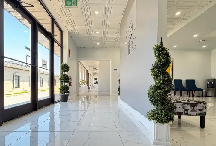 Bright modern lobby with white tiled floor, potted spiral plant, glass doors, and hallway with chairs.