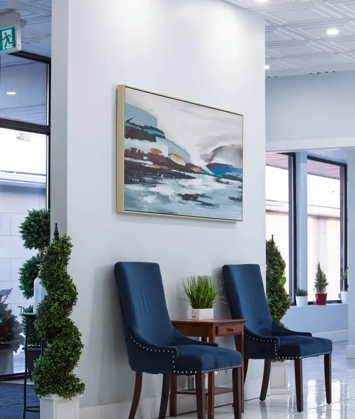 Two blue upholstered chairs with nailhead trim flank a small wooden table with a green plant.