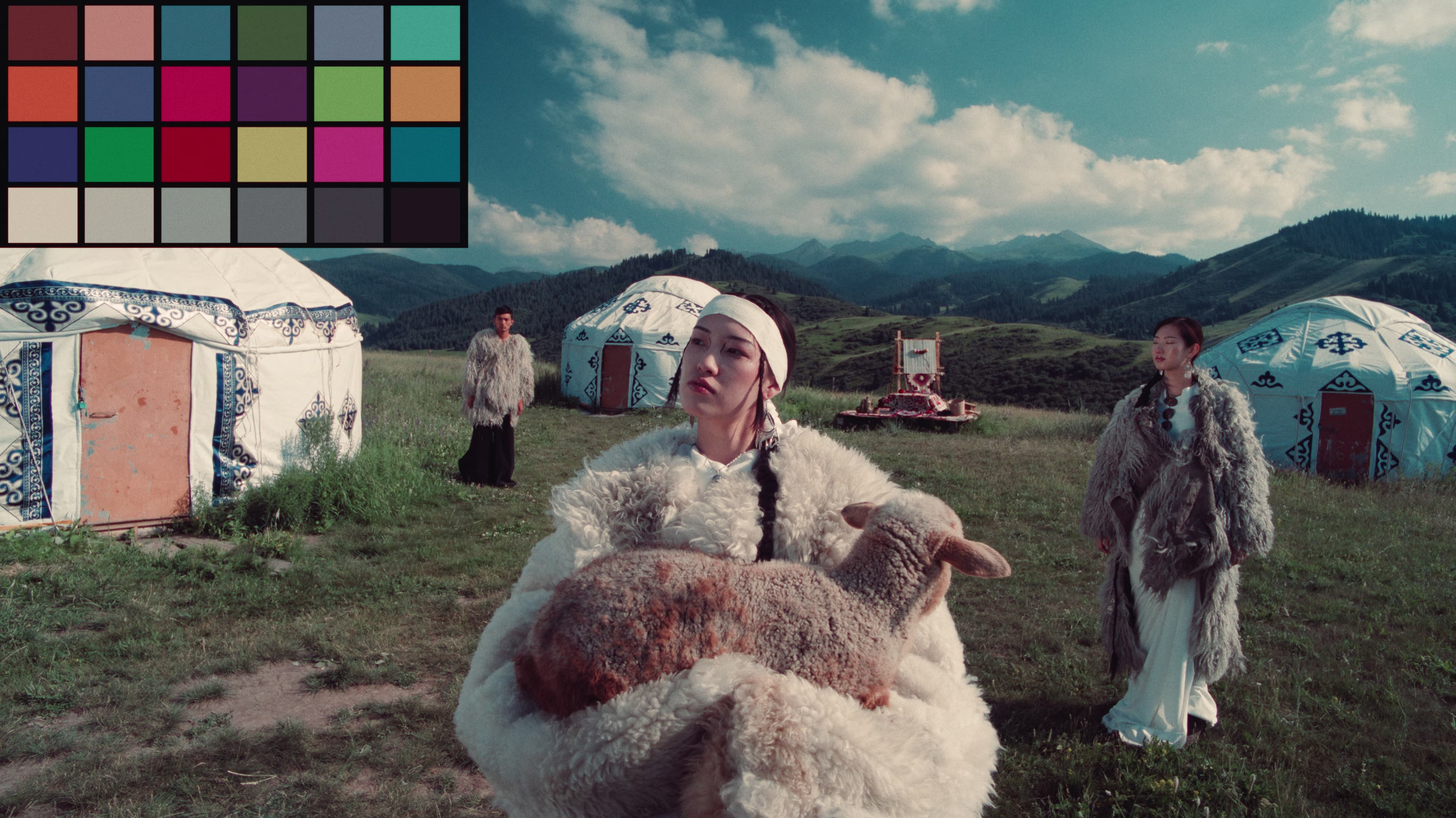 Three people in traditional fur clothing standing on grass with yurts and mountains under a partly cloudy sky; one person holds a sheep.