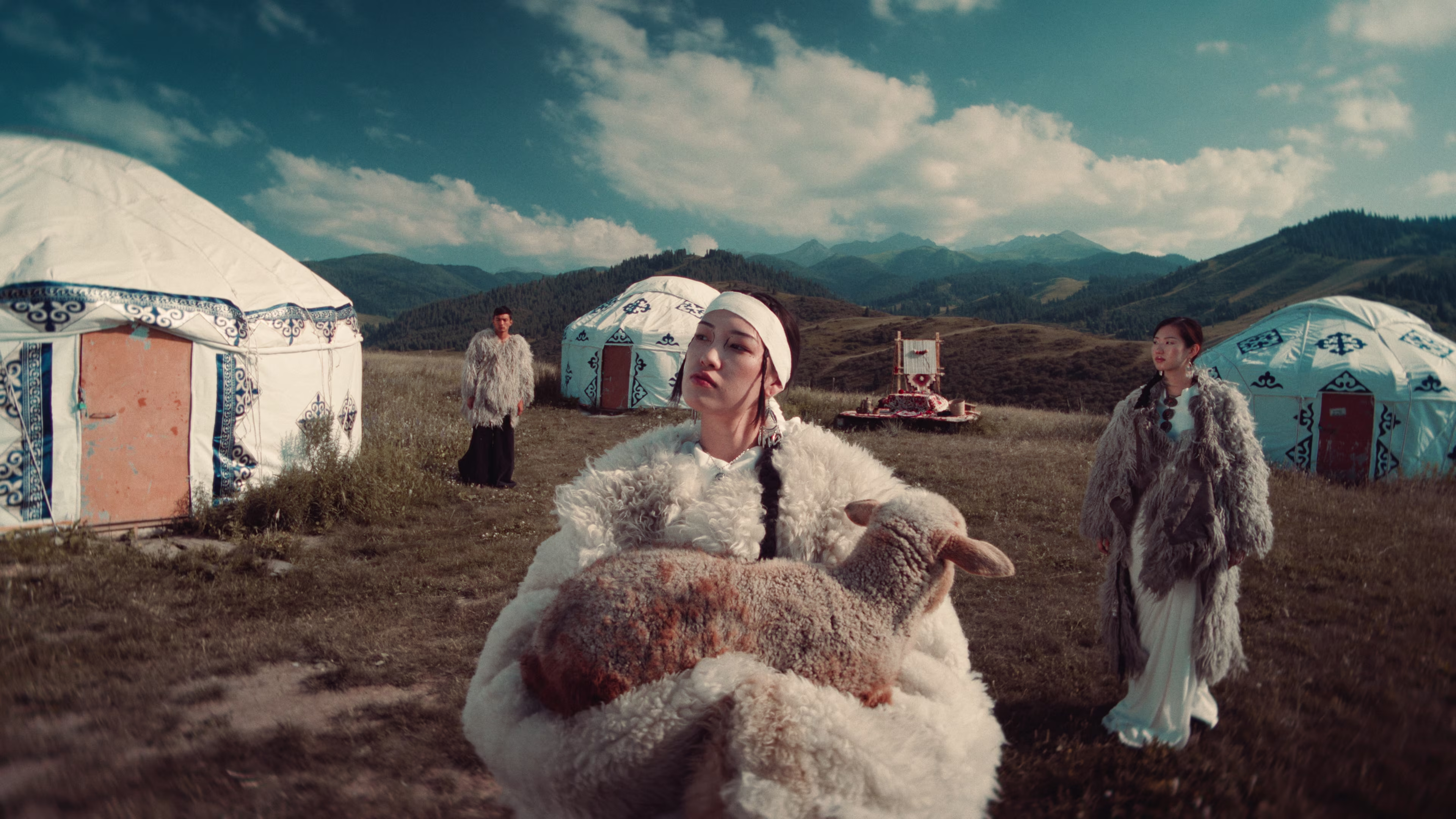 Three people in traditional fur clothing standing near decorated yurts in a grassy mountainous area, one person holding a lamb.