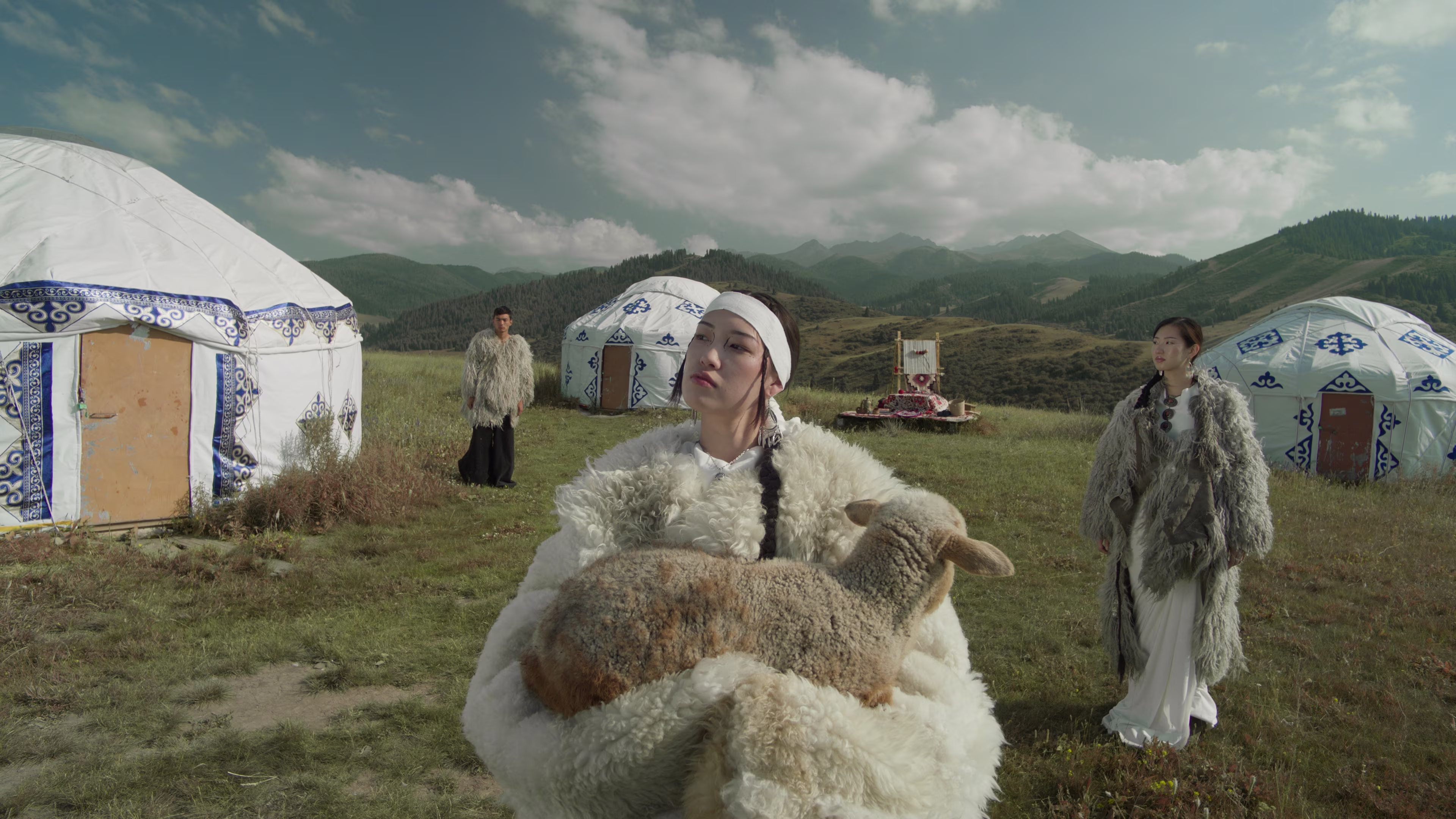 Three people in traditional fur clothing stand in a grassy field with white yurts and mountains in the background; the person in front holds a sheep.