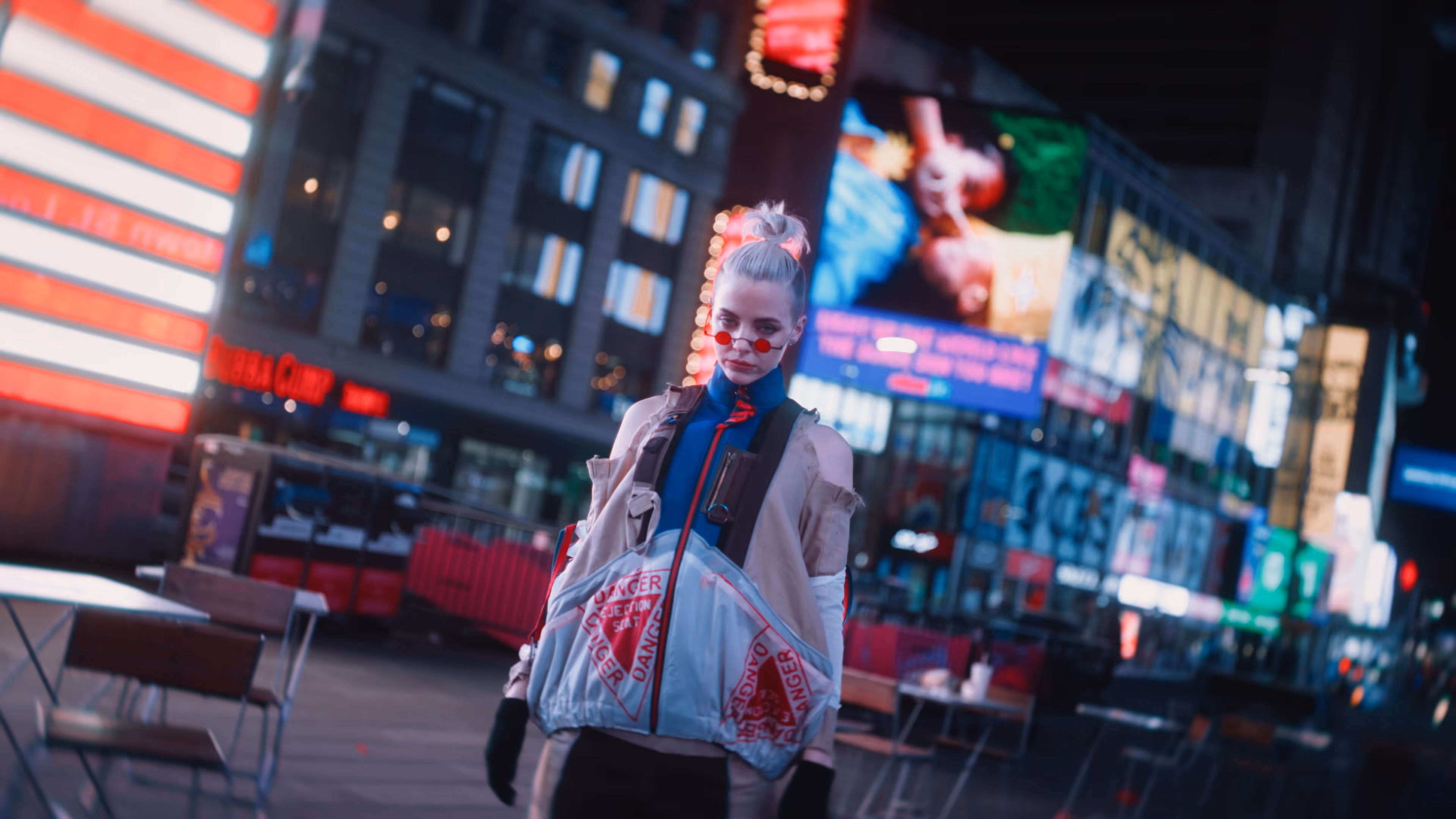 Person with light hair in a top bun wearing red-tinted glasses and a jacket with red 'DANGER' warnings, standing in a brightly lit urban area at night.