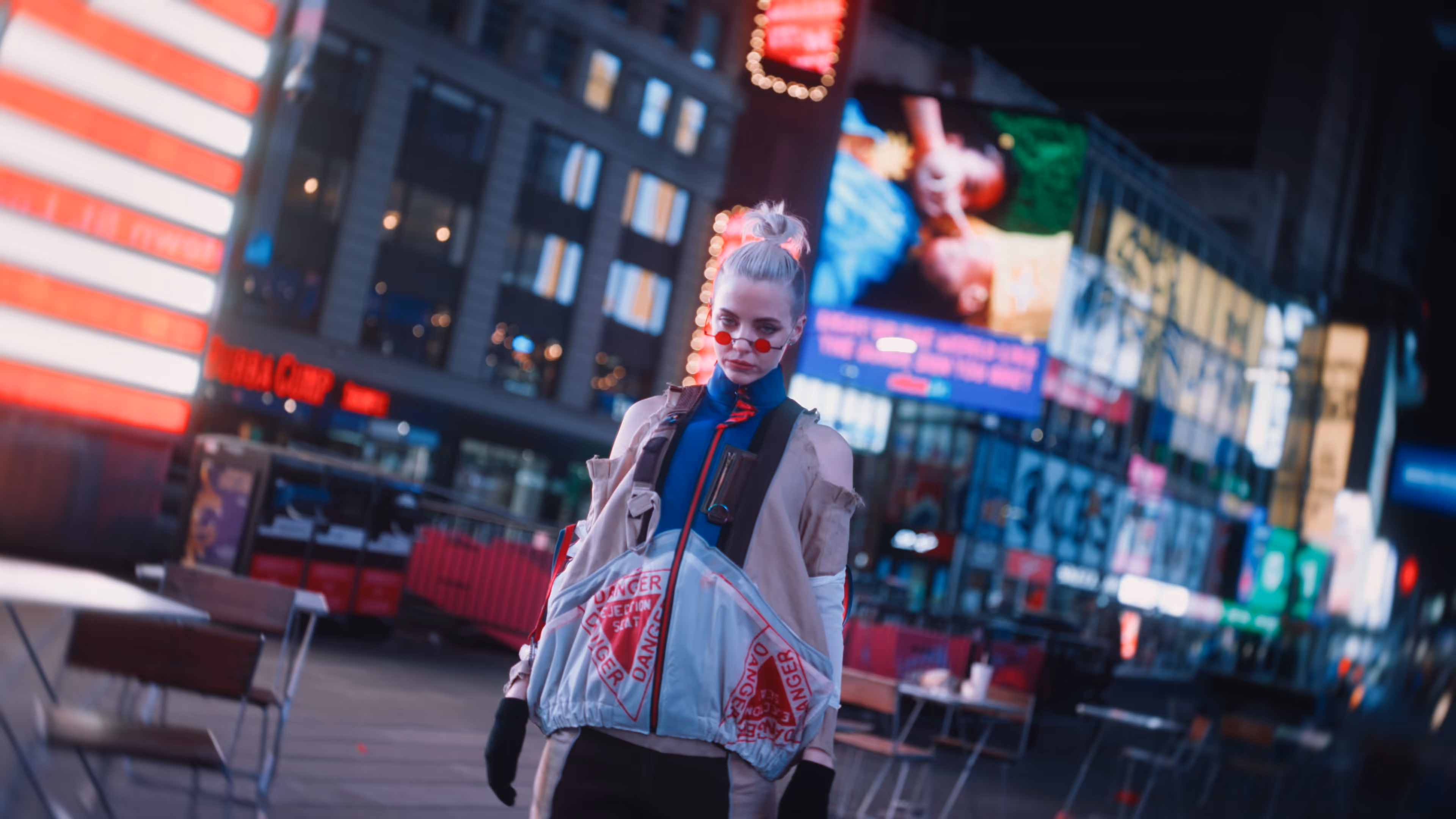 Young person with pale blonde hair in a bun and small red sunglasses standing in a city square at night with bright neon signs.