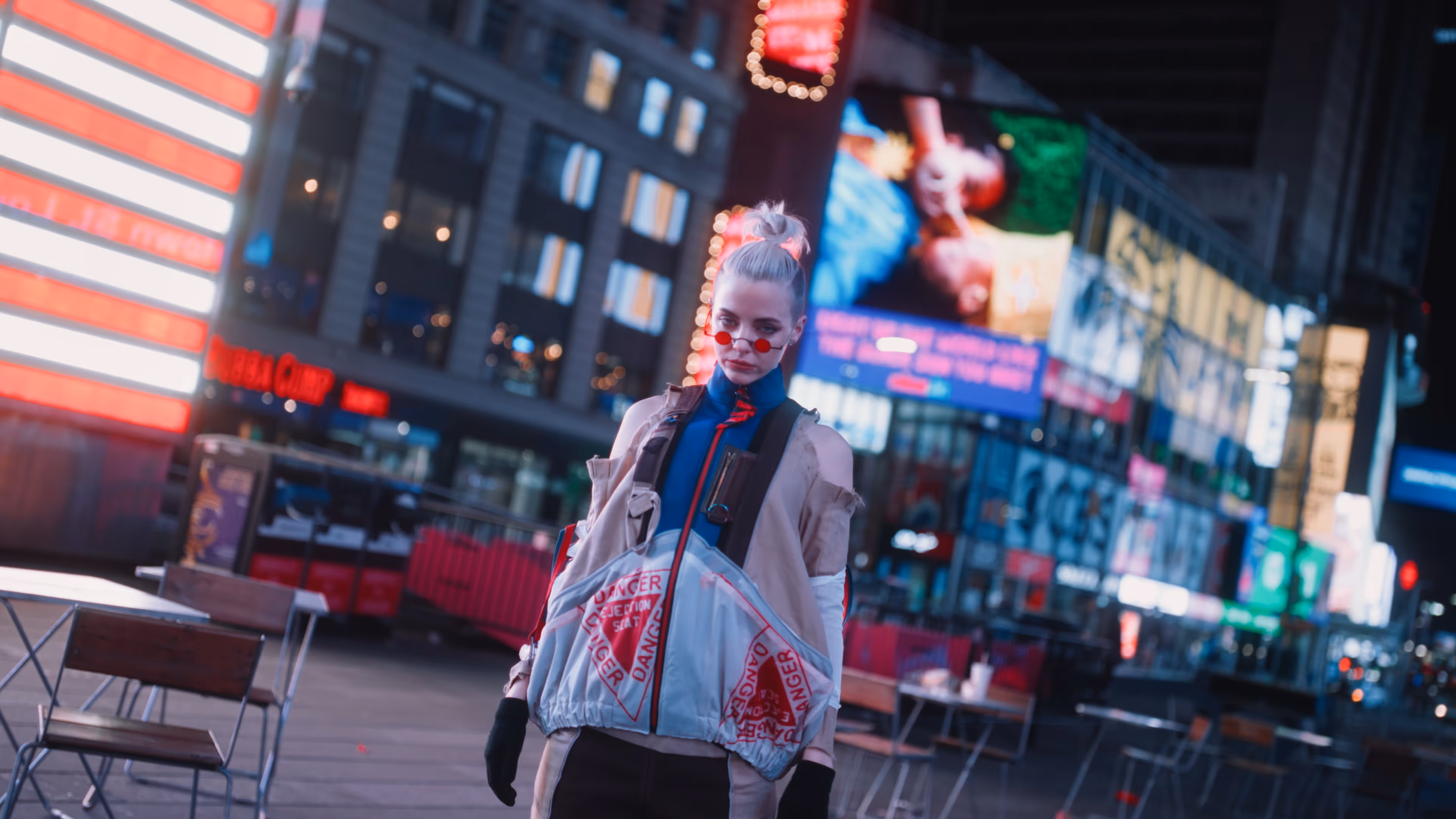 Person with light hair in a topknot, wearing red tinted glasses, a jacket with 'DANGER' printed pockets, and black gloves standing in a brightly lit urban area at night.