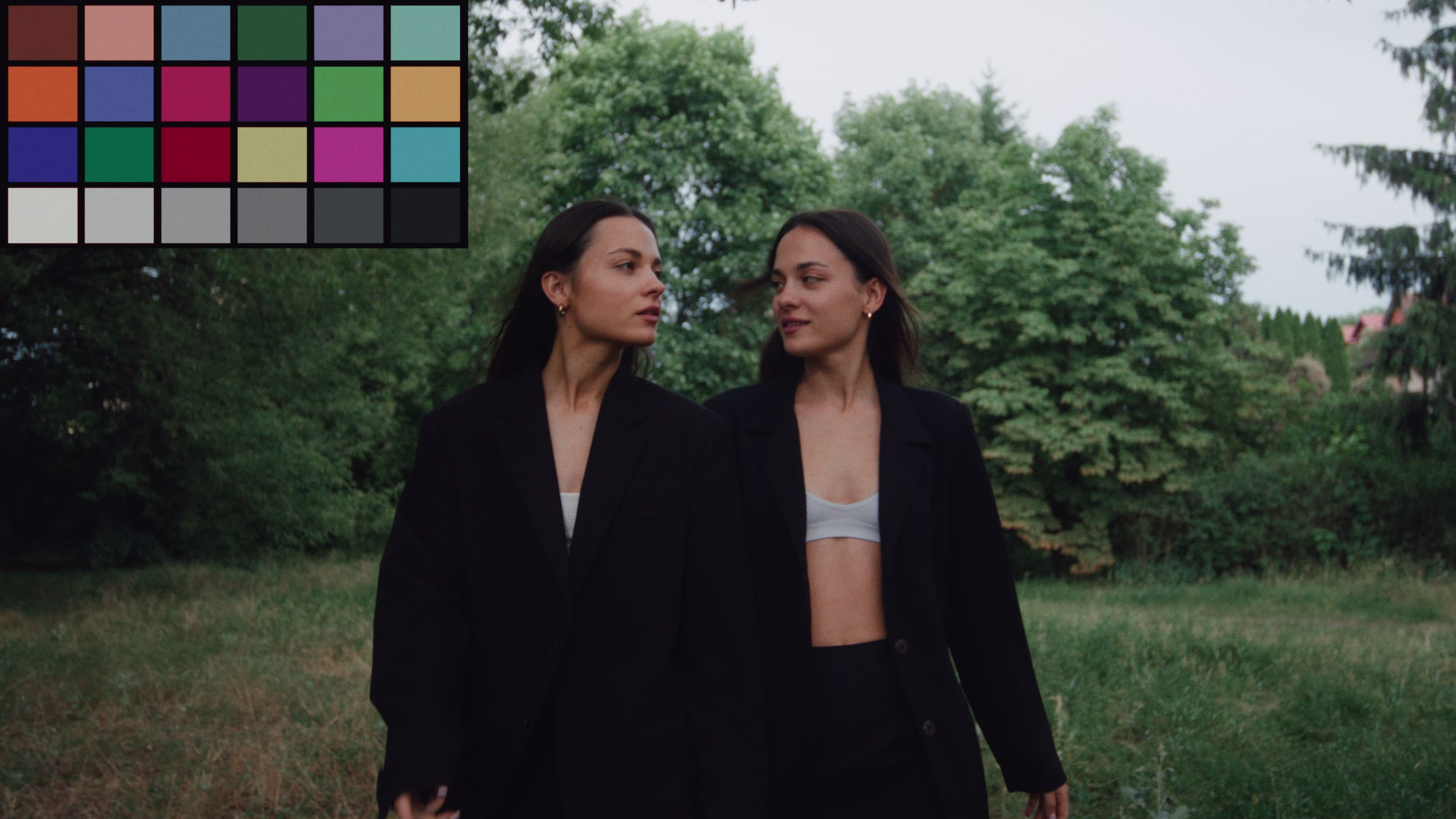 Two women wearing black outfits walking outside with green trees in the background, looking at each other.