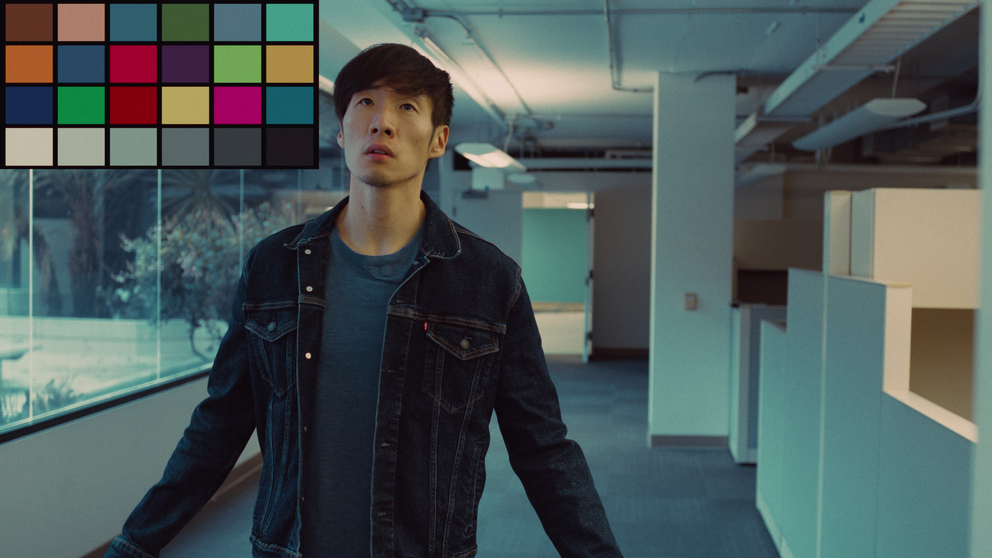 Man in a denim jacket standing in an office corridor with cubicles, looking upward thoughtfully.