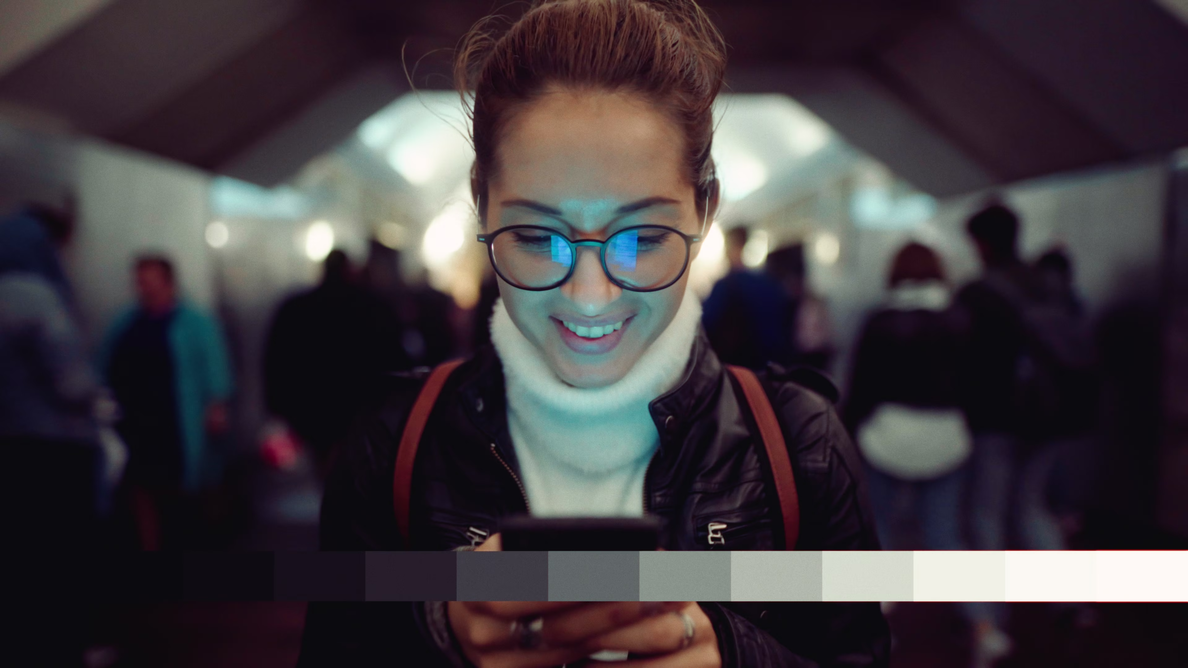Smiling woman wearing glasses and a turtleneck sweater looking at her smartphone in a busy indoor public space.