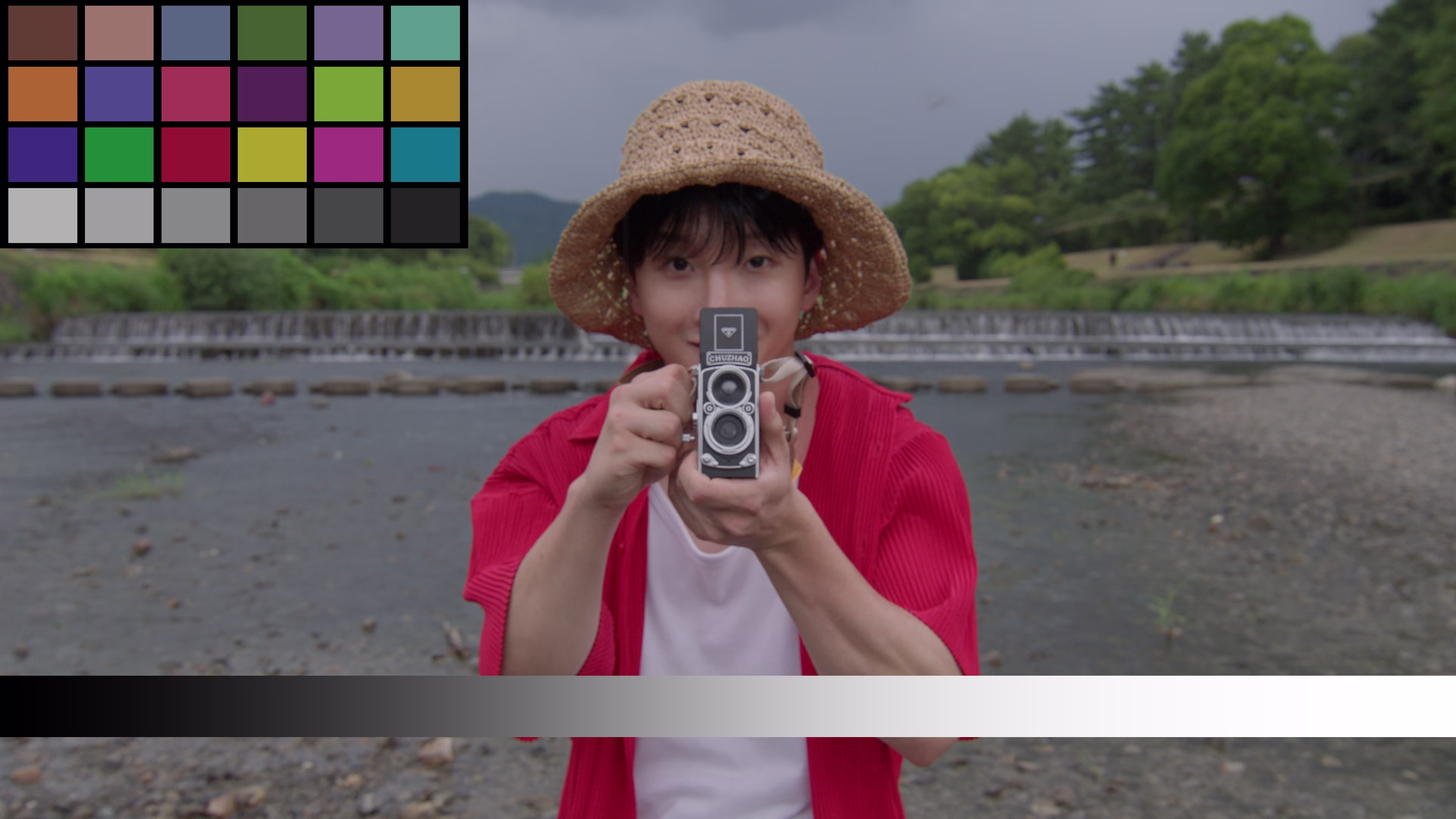 Person wearing a woven straw hat and red shirt holding a vintage twin-lens camera pointed at the viewer, standing near a river with greenery in the background.