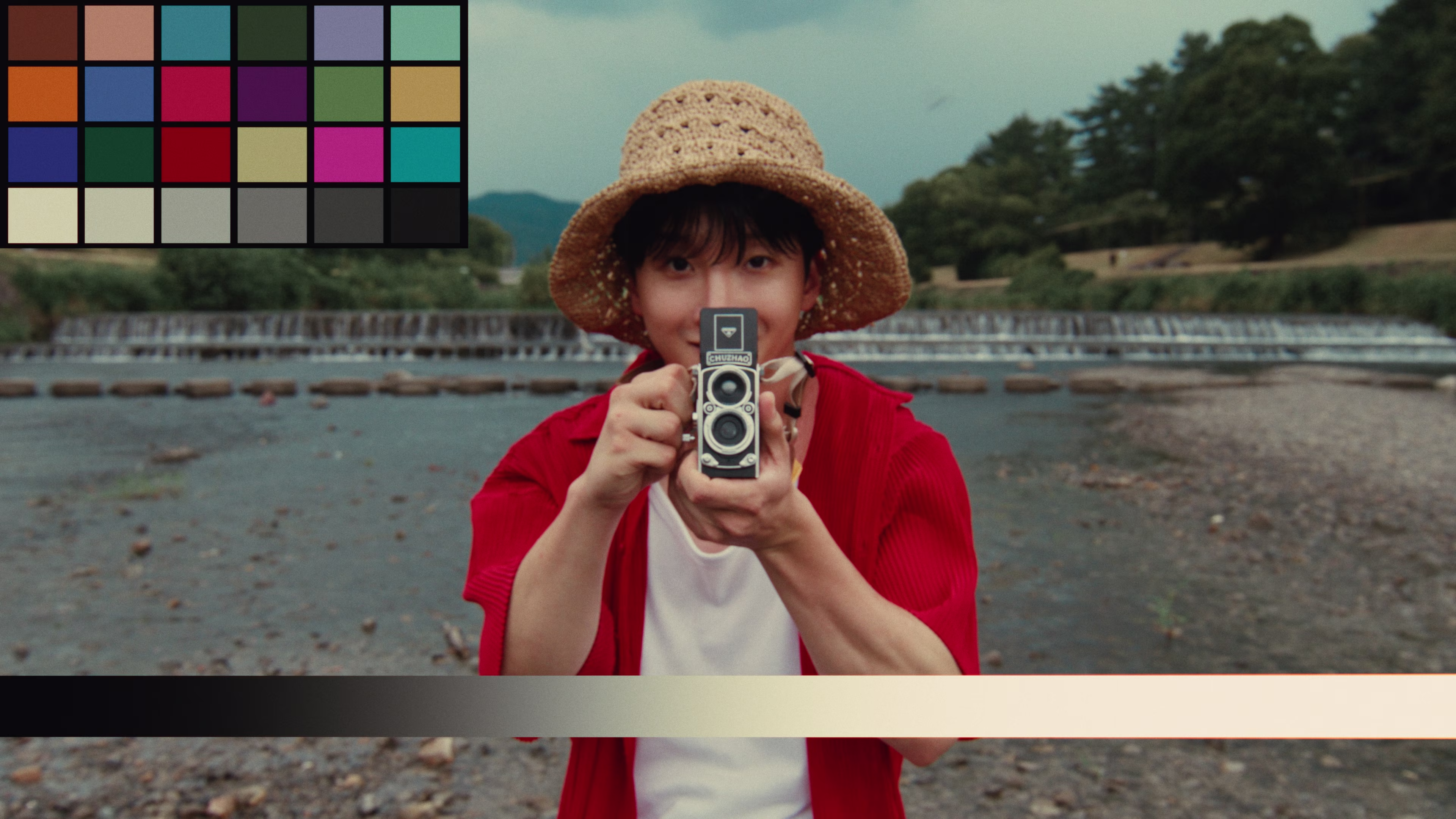 Person wearing a woven straw hat and red shirt holding a vintage twin-lens camera pointed at the viewer, standing near a river with greenery in the background.