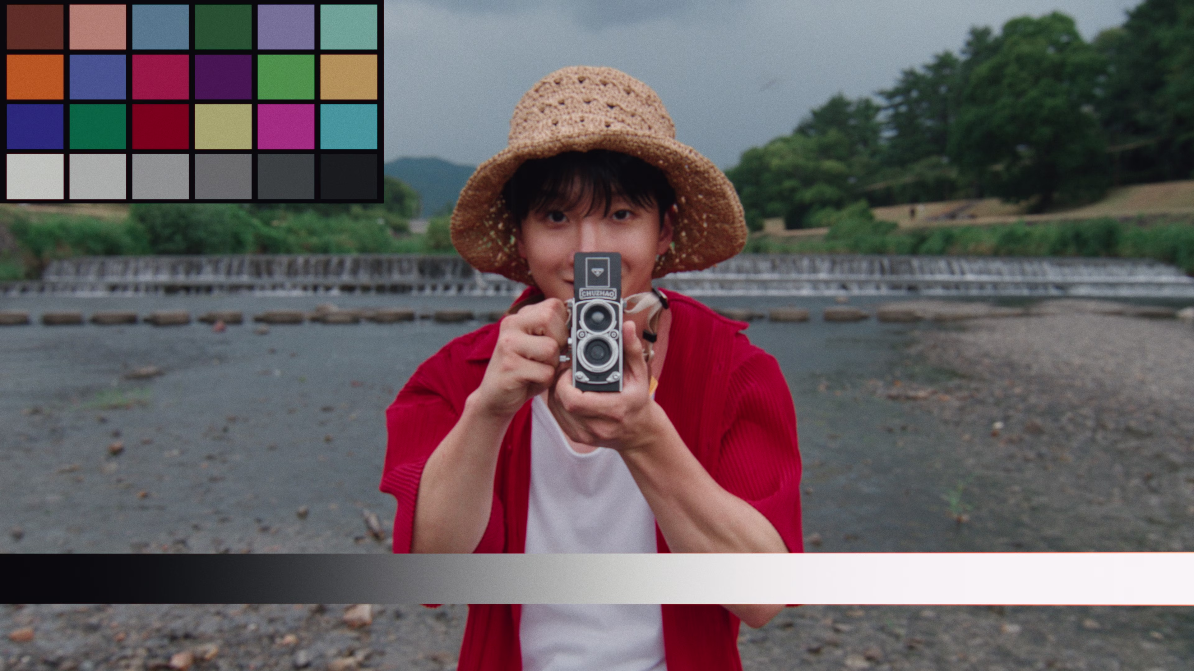 A person wearing a straw hat and red shirt holding a vintage twin-lens camera outdoors near a river with a small waterfall.