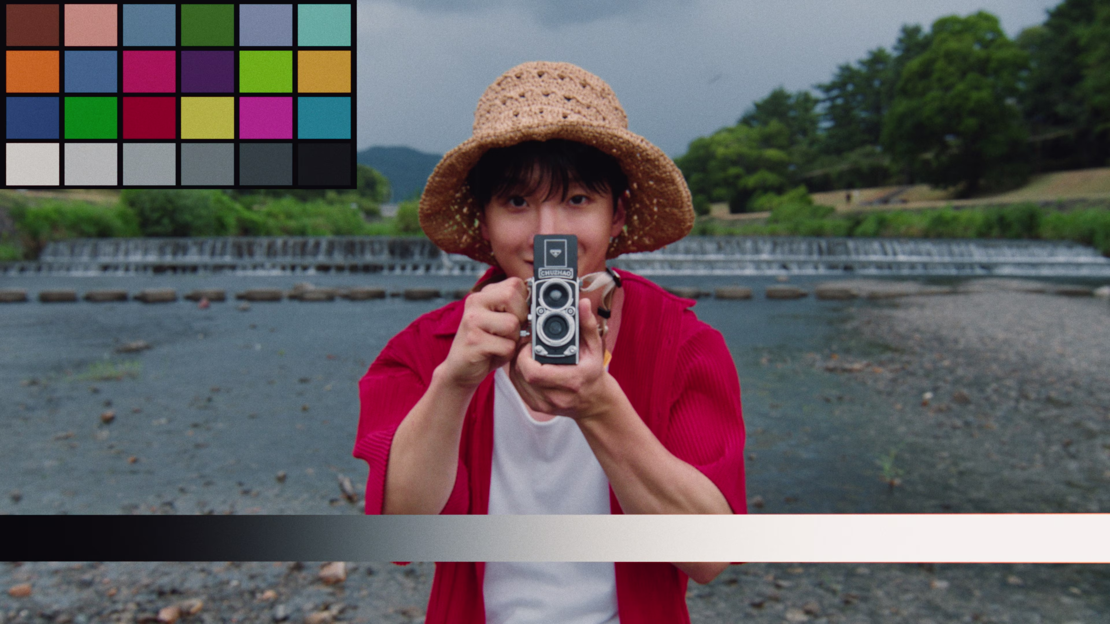 Person wearing a straw hat and red shirt holding a vintage twin-lens camera pointing towards the viewer, standing by a river with green trees in the background.