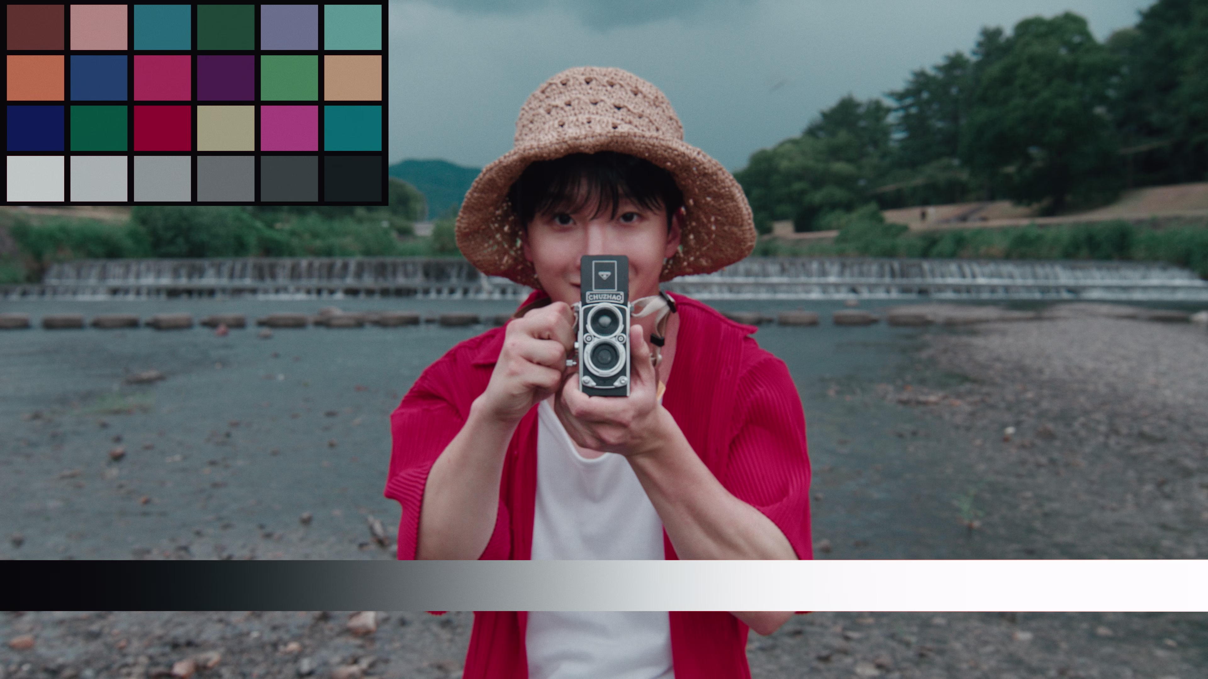 Person wearing a straw hat and red shirt holding a vintage twin-lens camera pointed at the viewer by a riverside.