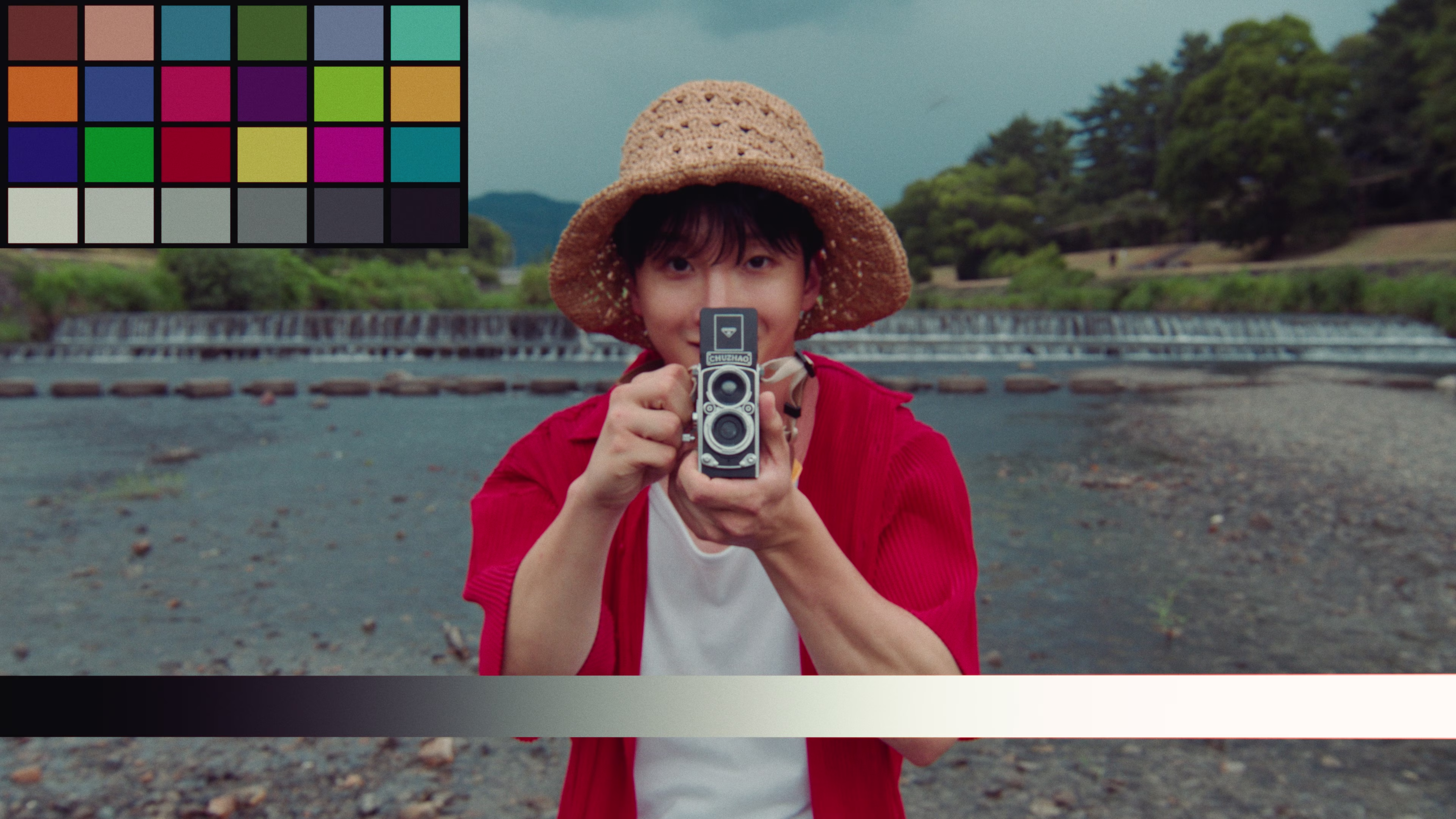 Person wearing a straw hat and red shirt holding a vintage camera pointed at the viewer, standing near a river with trees and a small waterfall in the background.