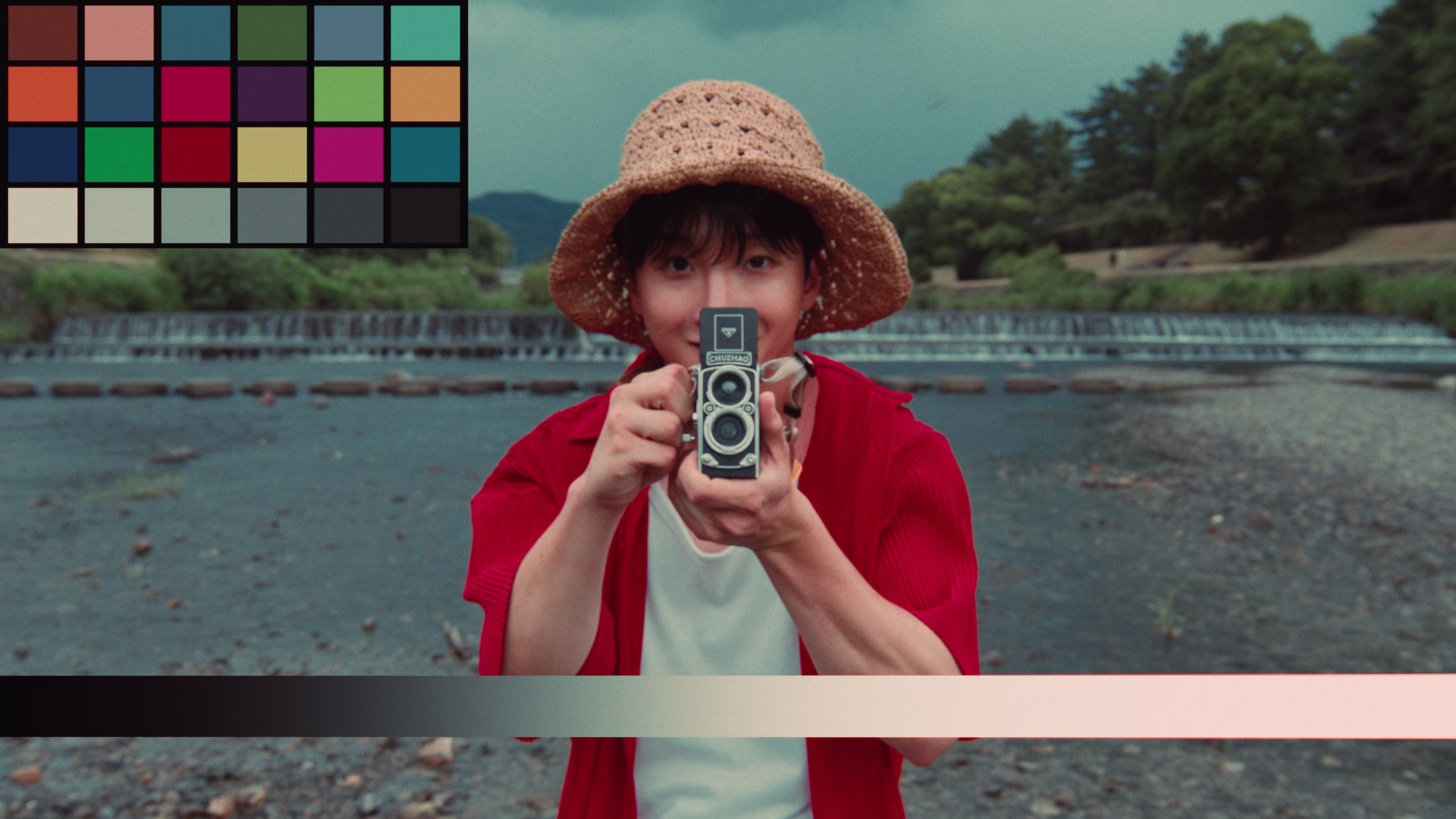 Person wearing a straw hat and red shirt holding a vintage twin-lens camera, standing outdoors near a river with trees and a waterfall in the background.