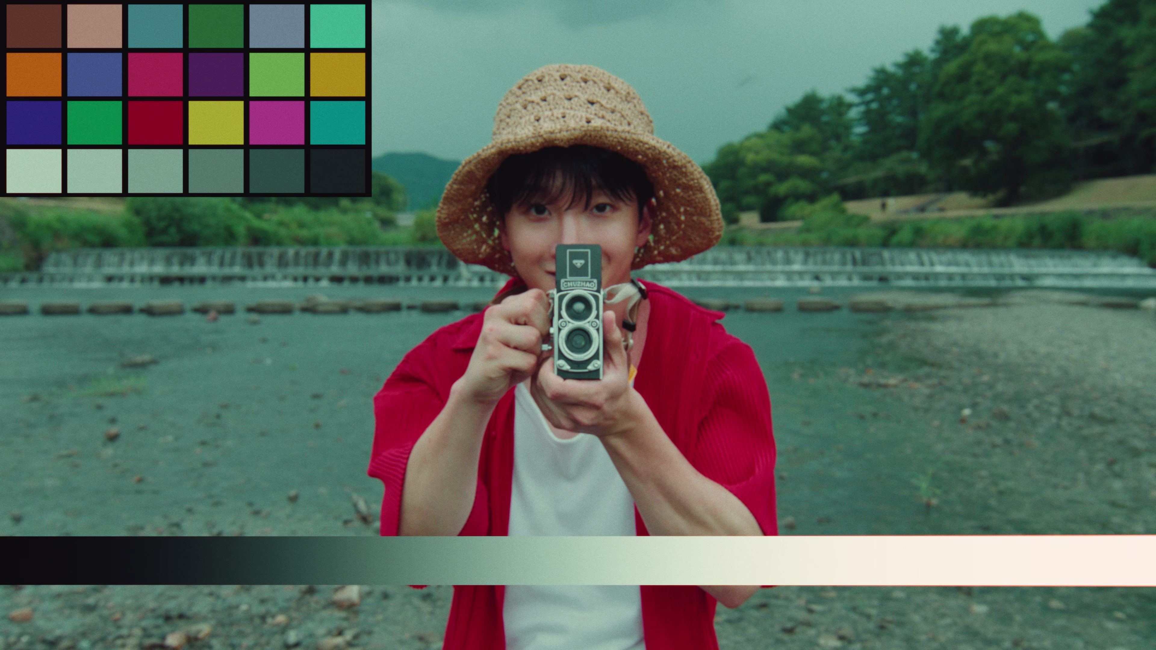 Person wearing a straw hat and red shirt holding a vintage twin-lens camera towards the viewer, standing near a river with trees in the background.