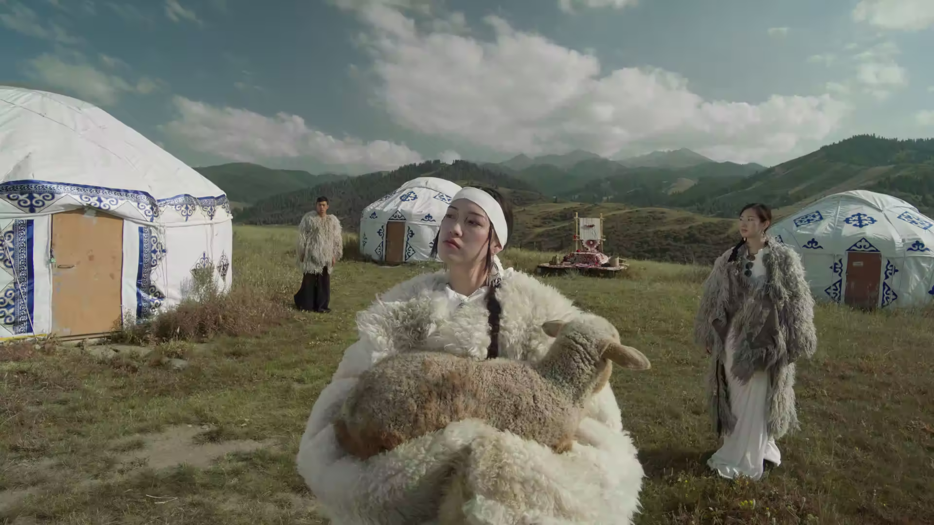 Three people in traditional fur clothing stand in a grassy field with white yurts and mountains in the background; the person in front holds a sheep.