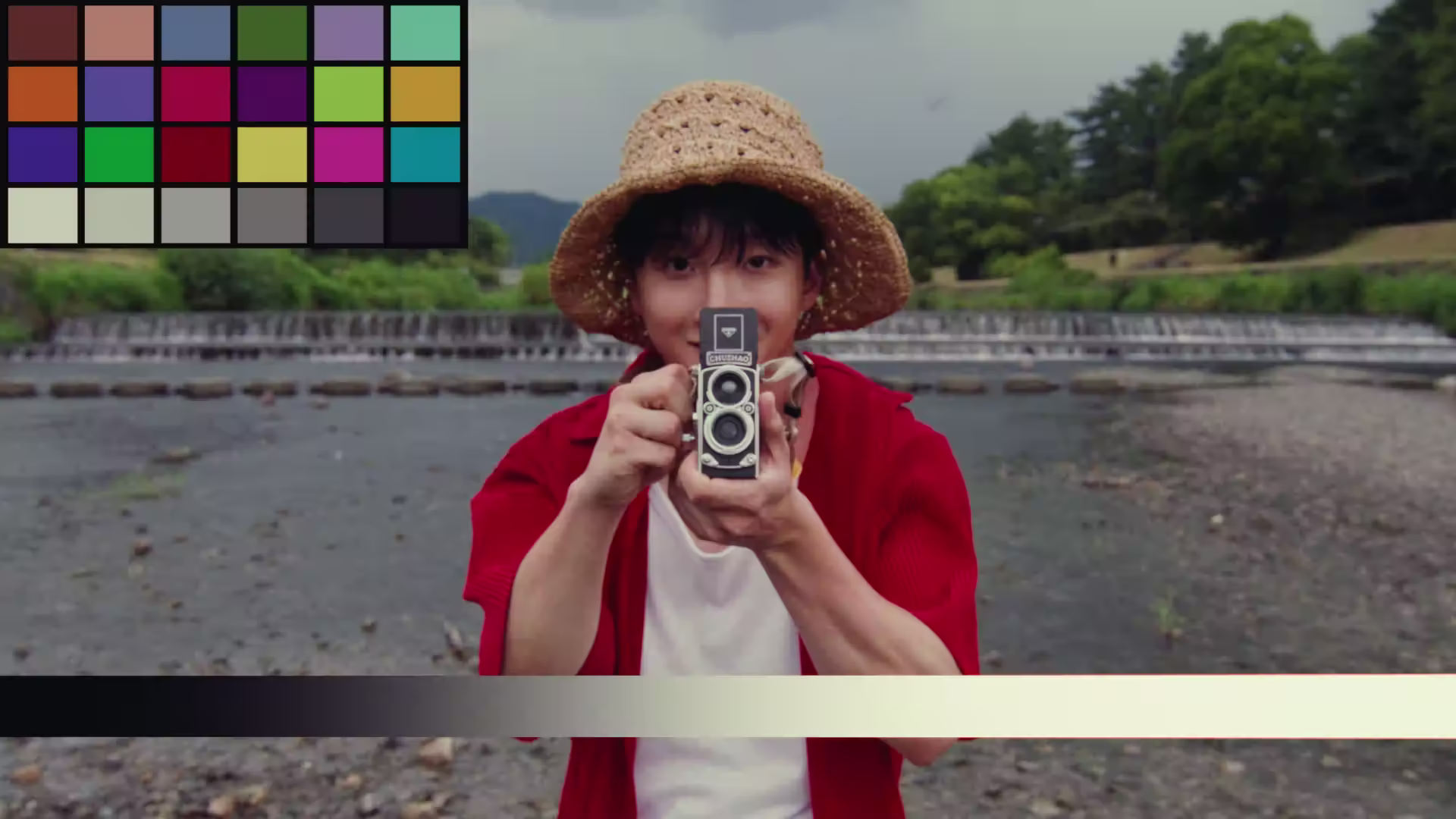 Person wearing a straw hat and red shirt holding a vintage twin-lens camera toward the viewer by a riverbank.
