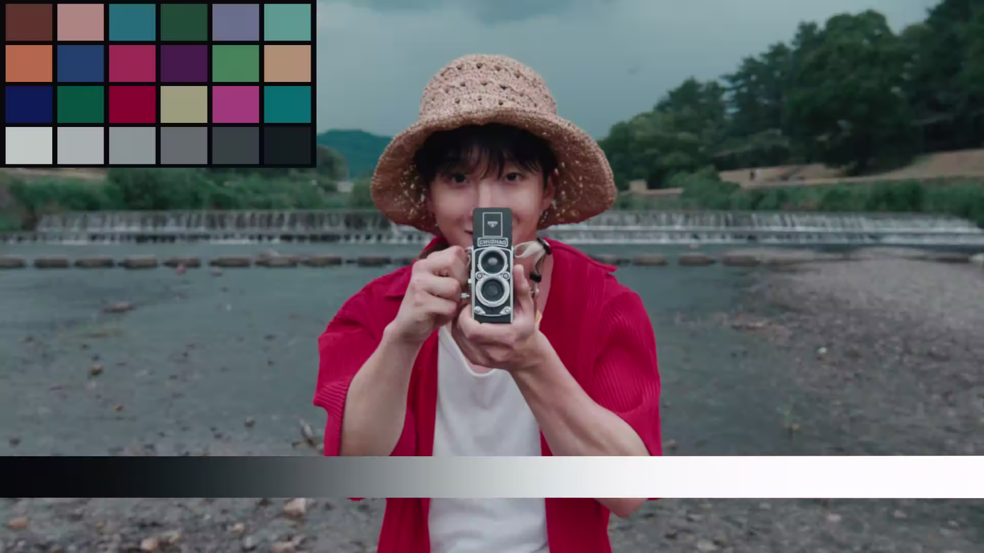 Person wearing a straw hat and red shirt holding a vintage twin-lens camera pointed at the viewer by a riverside.