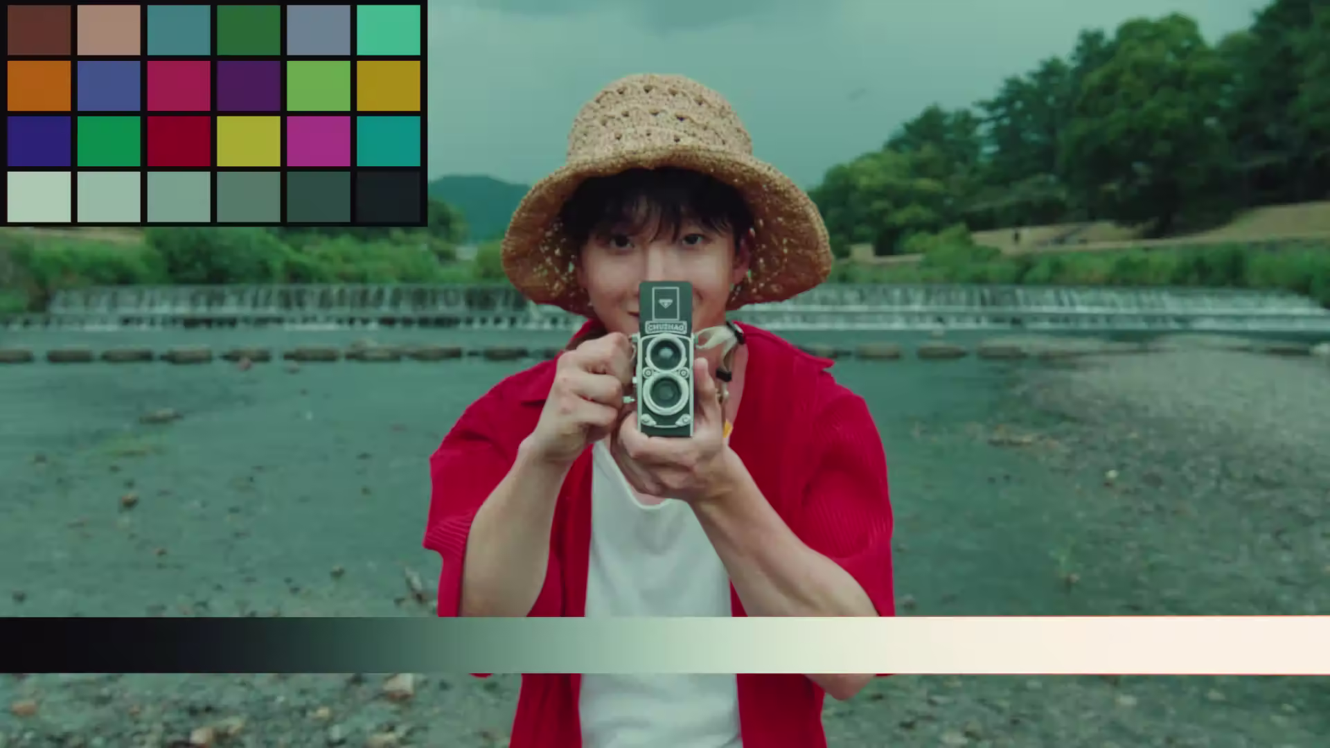 Person wearing a straw hat and red shirt holding a vintage twin-lens camera towards the viewer, standing near a river with trees in the background.