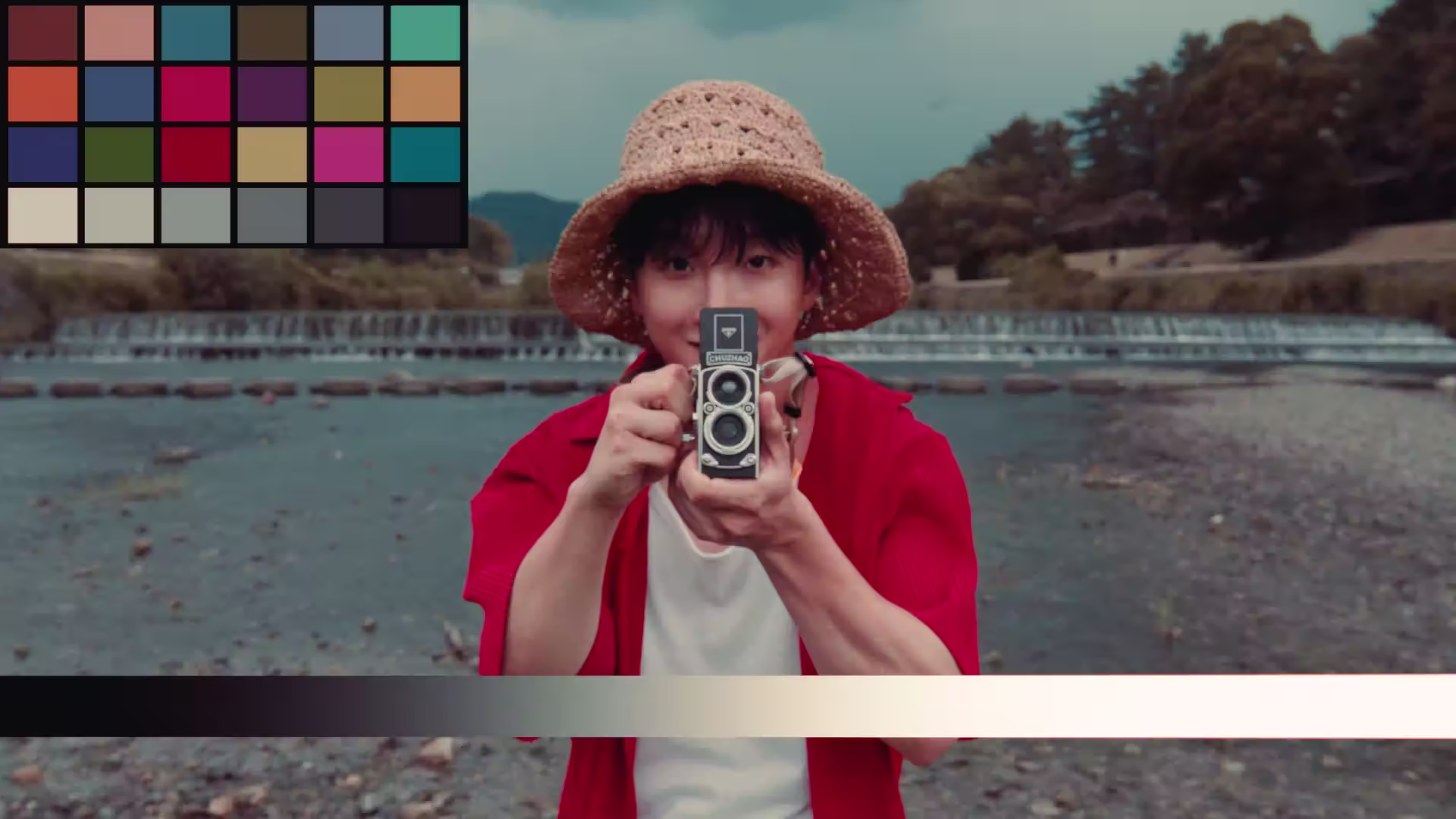 Person wearing a straw hat and red shirt holding a vintage twin-lens camera pointed at the viewer by a riverside with trees in the background.