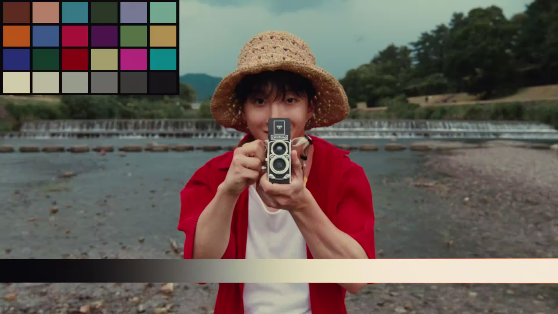 Person wearing a woven straw hat and red shirt holding a vintage twin-lens camera pointed at the viewer, standing near a river with greenery in the background.